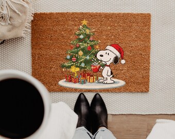 May include: A rectangular brown coir doormat featuring a printed illustration of Snoopy wearing a red Santa hat and holding a red mug, standing next to a decorated Christmas tree with colorful ornaments and wrapped gift boxes underneath.
