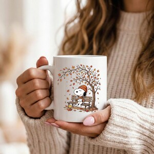May include: A white ceramic mug featuring a printed illustration of Snoopy wearing an orange beanie and scarf, sitting on a wooden bench under an autumn tree with falling orange and brown leaves, while holding a warm beverage in a cup.