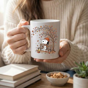 May include: A white ceramic mug featuring a printed illustration of Snoopy wearing an orange beanie and scarf, sitting on a wooden bench under an autumn tree with falling leaves, holding a cup while a small yellow bird sits nearby on the ground.