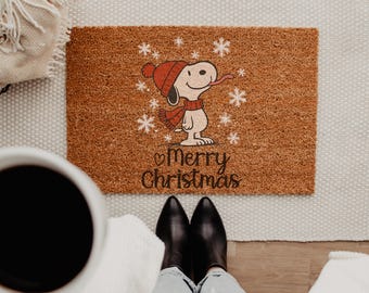 May include: A rectangular brown coir doormat featuring a graphic of Snoopy wearing a red knit hat and scarf, surrounded by white snowflakes. Below the character, the text Merry Christmas is printed in a dark brown cursive font.