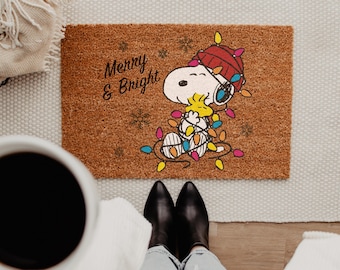 May include: A rectangular brown coir doormat featuring a graphic of Snoopy wearing a red winter hat and tangled in colorful Christmas lights while holding Woodstock. The text Merry & Bright is printed in black script with small snowflake icons.