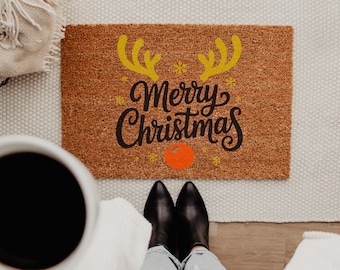 May include: A rectangular brown coir doormat featuring the black text Merry Christmas, yellow reindeer antlers, small yellow snowflakes, and a bright orange circular nose centered below the text on the mat surface.