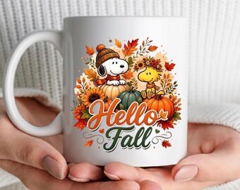 May include: A white ceramic mug featuring a colorful graphic of a cartoon dog wearing a knit hat and a yellow bird surrounded by orange pumpkins, sunflowers, and autumn leaves. The text Hello Fall is printed in orange and dark green script.