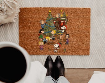 May include: A rectangular brown coir doormat featuring a colorful illustration of the Peanuts characters decorating a Christmas tree. Snoopy, Charlie Brown, Lucy, Linus, and other characters are shown in a festive holiday scene.