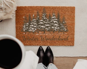May include: A rectangular brown coir doormat featuring a printed graphic of a forest of pine trees with white snow on the branches. The text "Winter Wonderland" is printed in a dark script font below the trees on the mat surface.
