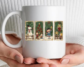 May include: A white ceramic mug featuring four vertical rectangular panels with Peanuts characters in winter scenes. The panels depict Snoopy with a snowman, Charlie Brown and Linus with a Christmas tree, Lucy ice skating, and Linus by a lamp post.