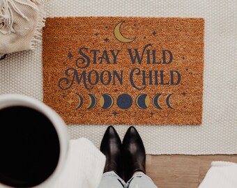 May include: A rectangular brown coir doormat featuring the text STAY WILD MOON CHILD in dark blue lettering. The design includes a yellow crescent moon, small stars, and a horizontal row of moon phases in blue and yellow tones.