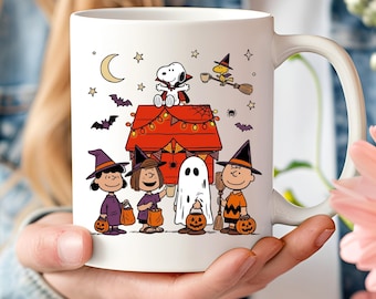 May include: A white ceramic mug featuring a Peanuts Halloween illustration. Snoopy sits on a red doghouse roof as a vampire, while Woodstock flies on a broom. Below, Lucy, Sally, Linus as a ghost, and Charlie Brown wear costumes and carry pumpkins.