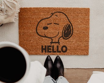 May include: A rectangular brown coir doormat featuring a black line drawing of the character Snoopy and the word HELLO printed in bold black capital letters centered below the illustration of the dog's head.