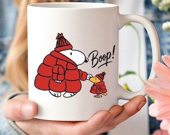 May include: A white ceramic mug featuring a graphic of a white dog and a yellow bird, both wearing red winter hats and red puffer jackets. The dog is touching the bird's nose with a finger, and the text Boop! is printed in black script next to them.