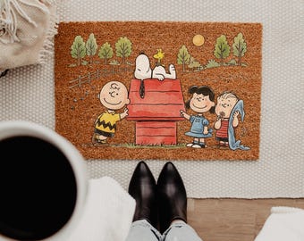 May include: A rectangular coir doormat featuring a colorful illustration of Charlie Brown, Snoopy, Woodstock, Lucy, and Linus standing by a red doghouse. The background shows a green landscape with trees, a fence, and a yellow sun in the sky.