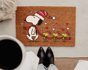 May include: A rectangular brown coir doormat featuring a graphic of Snoopy wearing a red and pink striped winter hat and scarf, leading three yellow Woodstock birds also wearing matching striped hats, surrounded by small red, pink, and white hearts.