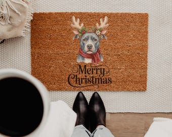 May include: A rectangular brown coir doormat featuring a central illustration of a gray pit bull wearing reindeer antlers and a red scarf. Below the dog, the text Merry Christmas is printed in a black, decorative, cursive script font.