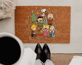 May include: A rectangular brown coir doormat featuring a colorful Peanuts holiday illustration. The graphic shows Snoopy, Charlie Brown, Lucy, Linus, Sally, and Woodstock surrounded by a Christmas tree, wrapped gifts, and string lights.
