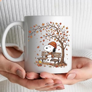 May include: A white ceramic mug featuring a graphic illustration of a white dog wearing an orange knit hat and scarf, sitting on a wooden park bench under a tree with falling autumn leaves, holding a coffee cup with a small yellow bird nearby.