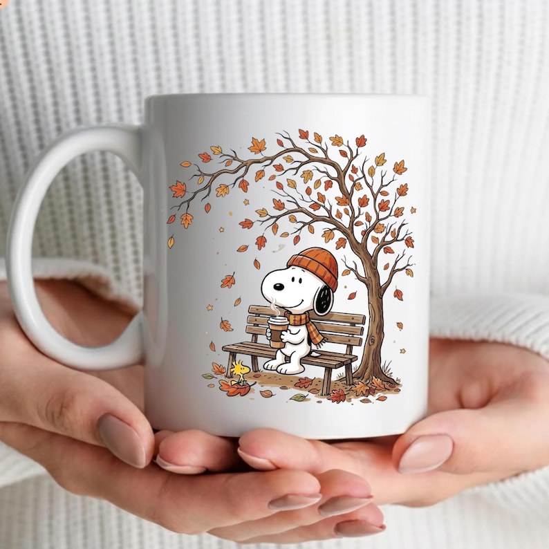 May include: A white ceramic mug featuring a graphic illustration of a white dog wearing an orange knit hat and scarf, sitting on a wooden park bench under a tree with falling autumn leaves, holding a coffee cup with a small yellow bird nearby.