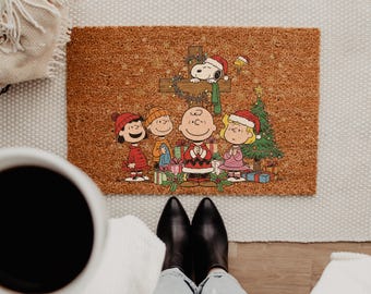 May include: A rectangular brown coir doormat featuring a Peanuts Christmas illustration. The design shows Charlie Brown, Snoopy, and friends gathered around a wooden cross and a decorated Christmas tree with colorful gift boxes.
