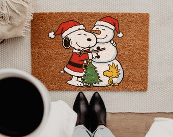 May include: A rectangular brown coir doormat featuring a printed illustration of Snoopy wearing a Santa hat and coat, hugging a snowman with a carrot nose and Santa hat, with Woodstock standing next to a small decorated Christmas tree.