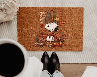 May include: A rectangular brown coir doormat featuring the word FALL in plaid and floral patterns. The design includes a cartoon Snoopy wearing a scarf, Woodstock, pumpkins, a basket, a lantern, mushrooms, apples, and autumn leaves.