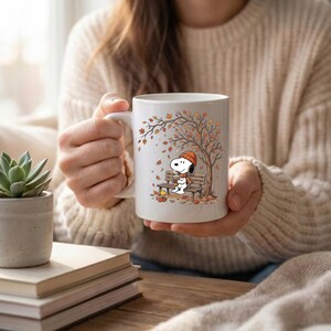 May include: A white ceramic mug featuring a printed illustration of Snoopy wearing an orange beanie and scarf, sitting on a wooden bench under an autumn tree with falling leaves, while holding a warm beverage with Woodstock nearby.