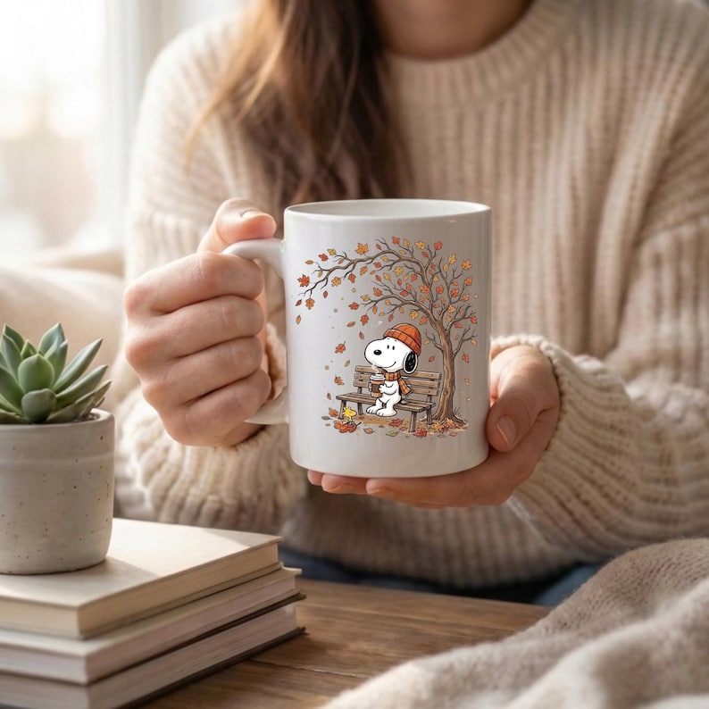 May include: A white ceramic mug featuring a printed illustration of Snoopy wearing an orange beanie and scarf, sitting on a wooden bench under an autumn tree with falling leaves, while holding a warm beverage with Woodstock nearby.