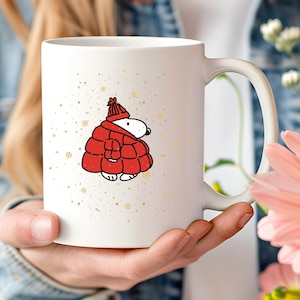 May include: A white ceramic mug featuring a graphic of a cartoon dog wearing a red puffer jacket and a matching red knit hat. The design is surrounded by small gold-colored star and snowflake accents scattered across the surface of the mug.