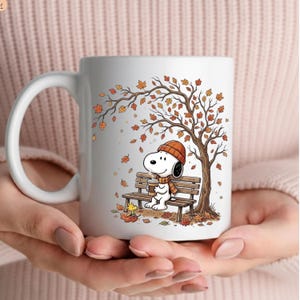 May include: A white ceramic mug featuring a printed illustration of Snoopy wearing an orange beanie and scarf, sitting on a wooden park bench under an autumn tree with falling orange and brown leaves, holding a coffee cup with a small yellow bird.