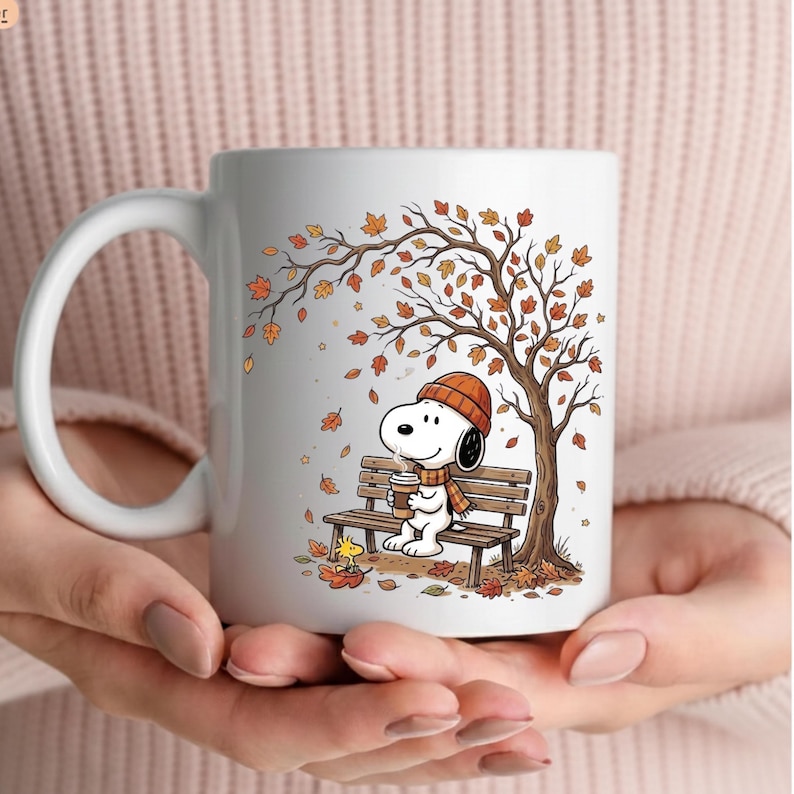 May include: A white ceramic mug featuring a printed illustration of Snoopy wearing an orange beanie and scarf, sitting on a wooden park bench under an autumn tree with falling orange and brown leaves, holding a coffee cup with a small yellow bird.