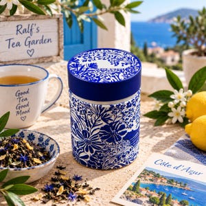 May include: A cylindrical tea canister featuring a deep blue and white floral pattern with the text Côte d'Azur on the lid. The canister is accompanied by a matching bowl filled with loose tea leaves and a mug with the text Good Tea Good Mood.