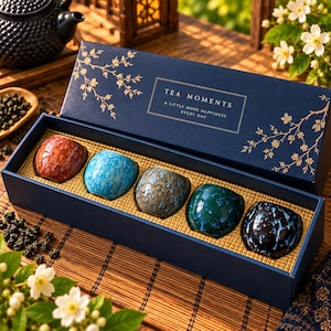 Könnte beinhalten: Eine rechteckige marineblaue Geschenkbox mit fünf kleinen Keramik-Teetassen mit glänzender, mehrfarbiger Craquelé-Glasur. Der Deckel ist mit goldenen Blumenranken und dem weißen Text "TEA MOMENTS A LITTLE MORE HAPPINESS EVERY DAY" verziert.
