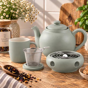 Könnte beinhalten: Eine salbeigrüne Keramik-Teekanne mit passender Tasse, ein Edelstahl-Teesieb mit grünem Sockel und ein runder Keramik-Stövchen mit Metallgitter. Ein Holzlöffel enthält losen Tee, daneben steht ein Glas mit Korkdeckel und der Aufschrift Tee.