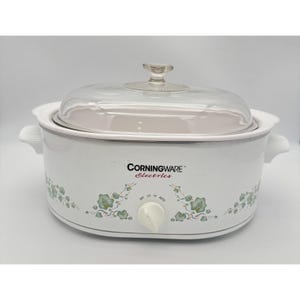 May include: A white oval CorningWare Electrics slow cooker with a clear glass lid. The exterior features a green and blue ivy leaf pattern, a central control knob with Off, Low, High, and Auto settings, and two side handles for lifting.