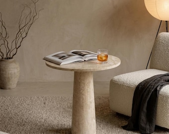 Op de afbeelding: Een ronde beige stenen bijzettafel met een taps toelopende voet. Op het tafelblad liggen een open boek en een glas met een amberkleurige vloeistof en ijsblokjes. De tafel heeft een natuurlijke, poreuze textuur en een neutrale kleurstelling.