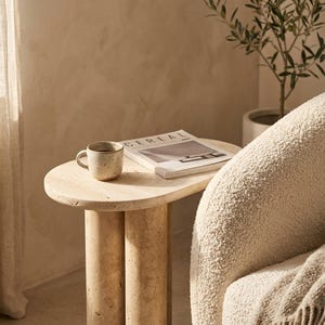 May include: A beige travertine side table with a smooth oval top and two cylindrical legs. A speckled ceramic mug and a magazine titled CEREAL are placed on the surface. The table is positioned next to a textured cream-colored armchair.