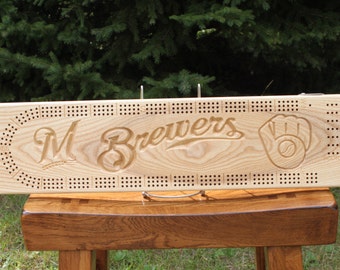 Könnte beinhalten: Ein hölzernes Cribbage-Brett mit dem Logo der Milwaukee Brewers, das in das hellfarbige Holz geschnitzt ist. Das Brett zeigt das "M"-Logo des Teams, das Wort "Brewers" und einen Baseballhandschuh. Das Brett hat Löcher für die Stifte.