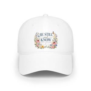 May include: A white baseball cap featuring a central floral wreath design with butterflies and the text BE STILL and KNOW THAT I AM GOD PSALM 46:10. The colourful botanical illustration includes pink, yellow, and blue flowers surrounding the lettering.