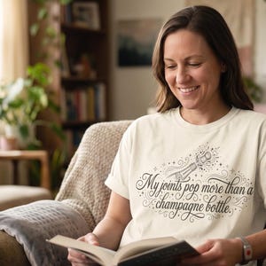 Könnte beinhalten: Ein cremefarbenes Kurzarm-T-Shirt mit einer dunkelbraunen Grafik einer knallenden Champagnerflasche und dem Text "My joints pop more than a champagne bottle." Das Design enthält dekorative Schnörkel und Sternakzente.