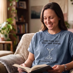 May include: A dusty blue short-sleeved t-shirt featuring a black graphic of an hourglass filled with spoons. The text reads: SAND RUNS, BUT I STILL HAVE SPOONS. MEASURING TIME IN ENERGY. The shirt has a crew neckline and rolled sleeve cuffs.