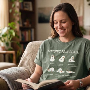 Puede incluir: Una camiseta de manga corta color verde salvia con un gráfico titulado CHRONIC PAIN SCALE. El diseño muestra seis ilustraciones de muñecos de nieve en varios estados de fusión, etiquetados desde No Pain hasta Worst Pain Possible, con texto.