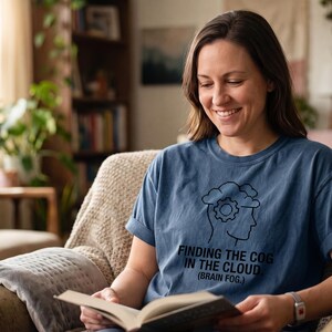 May include: A blue short-sleeved t-shirt featuring a black line art graphic of a human head silhouette with a gear inside a cloud. The text below the graphic reads: FINDING THE COG IN THE CLOUD. (BRAIN FOG.) The shirt has a crew neckline and rolled sleeves.