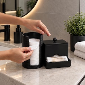 May include: A matte black cylindrical cotton pad dispenser with a vertical cutout and a matching square cotton swab holder featuring a small knob on the lid. The set is designed for bathroom organisation and holds white cotton rounds and swabs.