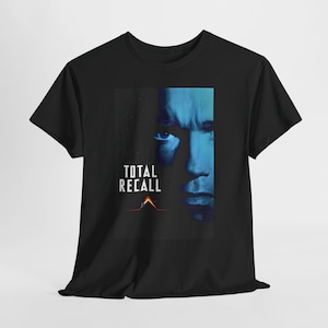 Könnte beinhalten: Ein schwarzes T-Shirt mit kurzen Ärmeln und einem Grafikdruck eines Gesichts in Blautönen auf der rechten Seite. Der Text "TOTAL RECALL" ist links in Weiß gedruckt, über einem kleinen dreieckigen Logo in Rot und Orange mit einer Linie.