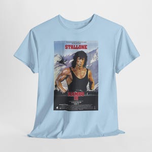 May include: A light blue short-sleeved t-shirt featuring a graphic print of the Rambo III film poster. The print displays Sylvester Stallone in a black tank top against a mountain backdrop, with red text reading STALLONE and RAMBO III.