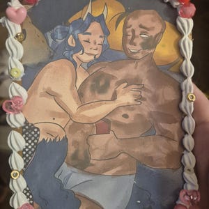 May include: A rectangular art print featuring an illustration of two figures embracing, framed by a border of white faux-whipped cream piping, pink heart-shaped beads, and small colorful circular accents. The illustration depicts a blue-haired figure with horns hugging a larger, dark-skinned figure. The overall aesthetic combines a hand-drawn character scene with decorative, textured craft elements.