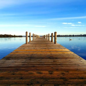 以下が含まれることがあります： 澄んだ空の下、穏やかな青い湖に長く伸びる木製の桟橋。水平に並んだ板と両側に立つ垂直の木の柱で構成された構造物が、木々のある遠くの岸辺に向かって続いています。
