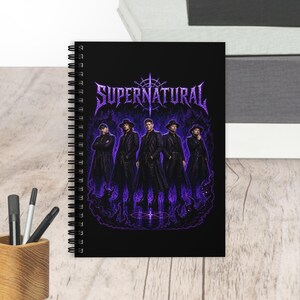 May include: A black spiral-bound notebook featuring a graphic print of the word SUPERNATURAL in purple lettering above five figures in long coats standing amidst purple flames and swirling smoke on a dark background.
