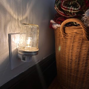 May include: A plug-in night light featuring a small, clear glass mason jar with embossed markings including the text 50 and 100. The jar is attached to a white electrical base with a metal ring, emitting a warm, glowing light from within.