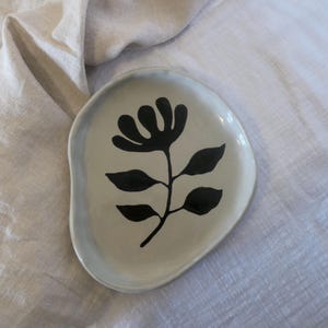 Pode incluir: Um prato de cerâmica com formato irregular e borda cinza claro. O prato apresenta um desenho floral preto no centro. O prato está sobre uma superfície de linho branco.