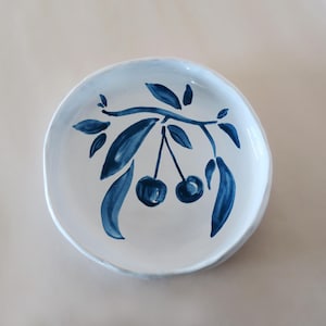 Puede incluir: Plato de cerámica blanco con un diseño pintado a mano de cerezas y hojas azul oscuro sobre una rama. El plato tiene una forma irregular y un borde azul claro. Ideal para servir porciones pequeñas.