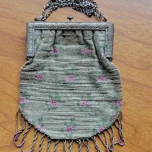 Può includere: Una piccola borsa di perline con una cornice in metallo argentato con un intricato motivo inciso. Il corpo è ricoperto da minuscole perline grigie con un motivo floreale ripetuto in rosa e verde. Una frangia di perline pende dal bordo inferiore.